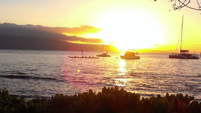 A 6 person outrigger canoe paddles past moored sailboats. The sunsets in the background.